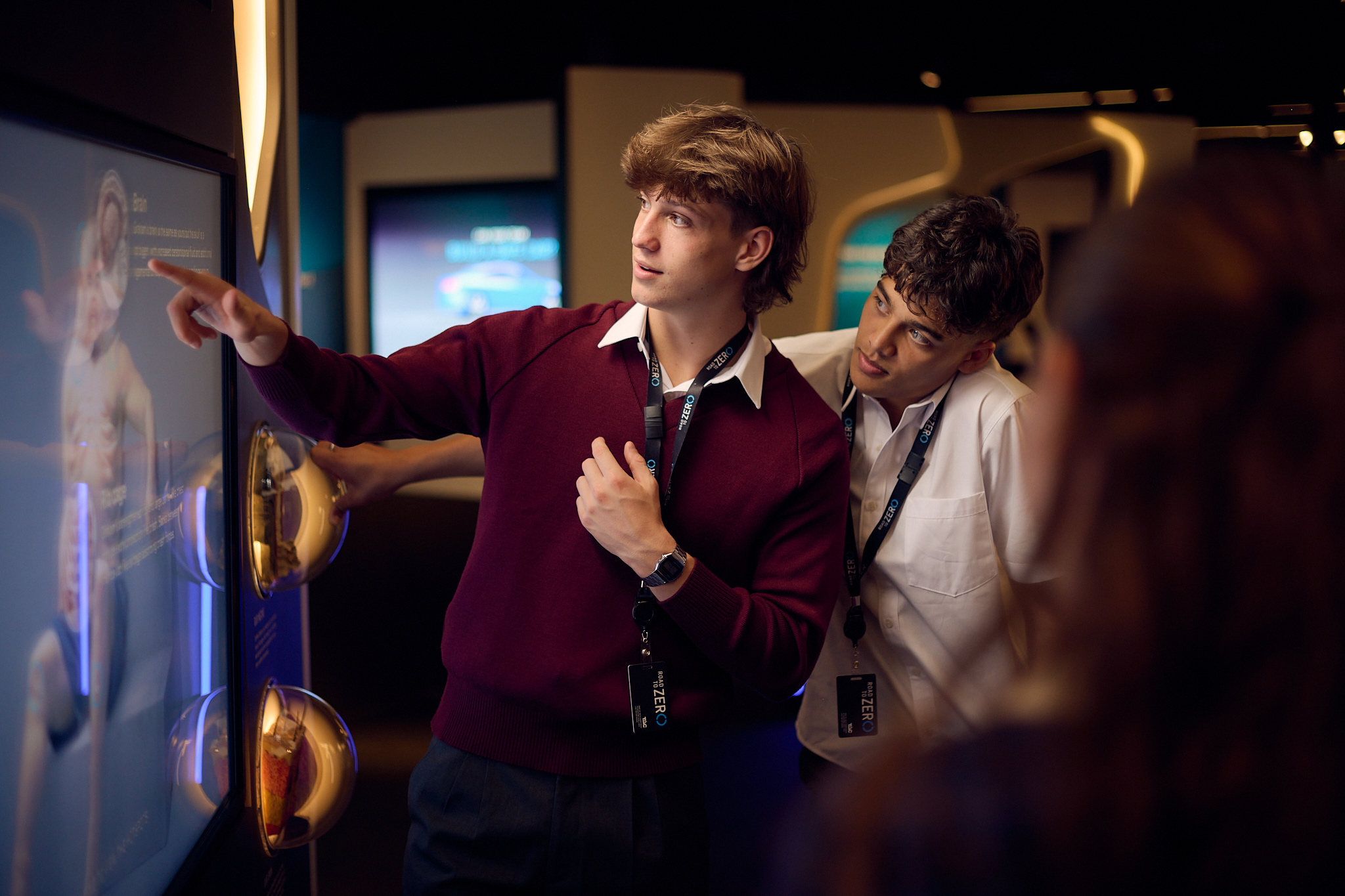 Two teenage boys in school uniform interact with a large wall screen